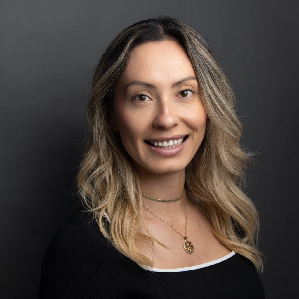 Professional headshot of a woman with shoulder-length wavy blonde hair, wearing a black top with a white trim and a necklace, smiling confidently in front of a neutral dark background.